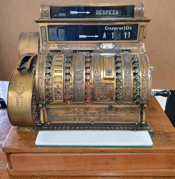 COLECIONISMO - NATIONAL - Rara maquina registradora confeccionada em bronze dourado de excepcional fundição e manufatura americana National, Fabricação 1914. Ricamente cinzelada e decorada com flores, acantos, palmas e volutas em relevo, sobre robusta base em madeira de lei, na parte inferior acompanha caixa. Funcionando todos os comandos. Excelente estado de conservação. Mede aprox. 56 x 62 x 40 cm. — COLECIONISMO
