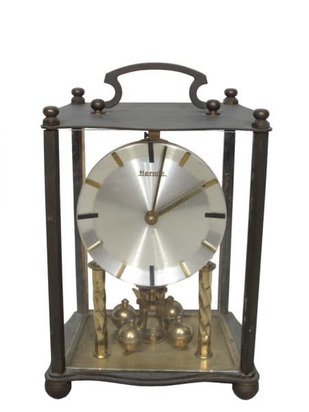 HERMLE (Alemanha, 1922), Relógio de mesa, estante ou cornija, 400 dias, com estrutura confeccionada em bronze, laterais envidraçadas, base e parte superior recortada com alça articulável, mostrador redondo em metal prateado, pêndulo giratório, faltando lateirais em vidro, faltando chave, ausência das laterais e traseira em vidro, não testado e sem garantia de funcionamento, medindo 22x14x11cm. — HERMLE