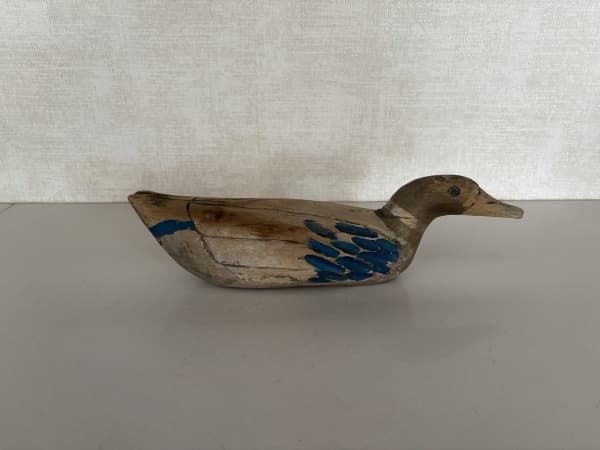 PATO DECOY DE MADEIRA ENTALHADA, (TONS DE AZUL E MARROM), ESTILO FOLK ART NORTE-AMERICANO, DESTACA-SE PELA AUTENTICIDADE, TEXTURA RÚSTICA E VALOR COMO PEÇA HISTÓRICA COLECIONÁVEL. APRESENTA DESGASTE VISÍVEL NA PINTURA ORIGINAL E RACHADURA NATURAL DA MADEIRA NO PESCOÇO. MEDIDAS APROXIMADAS: 33 CM X 9 CM X 9 CM — PATO DECOY DE MADEIRA ENTALHADA